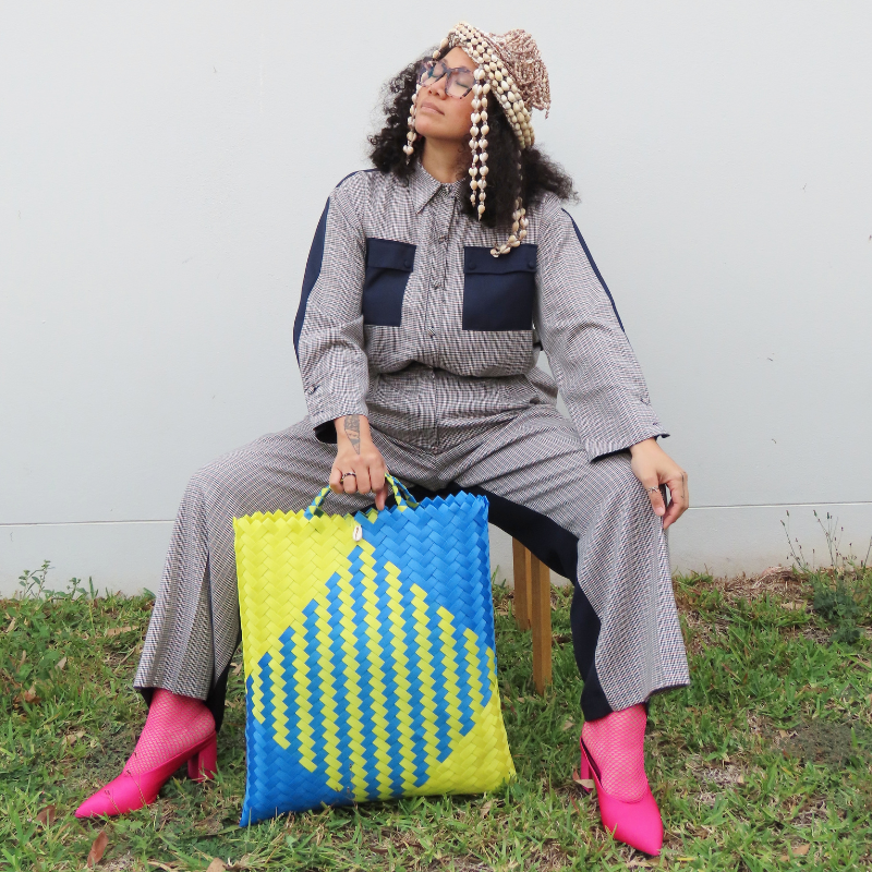 Maryann sitting on a stool on the grass, holding a woven yellow and blue bag. She is weaving a suit and shell headpiece, pink fishnet socks and heels.
