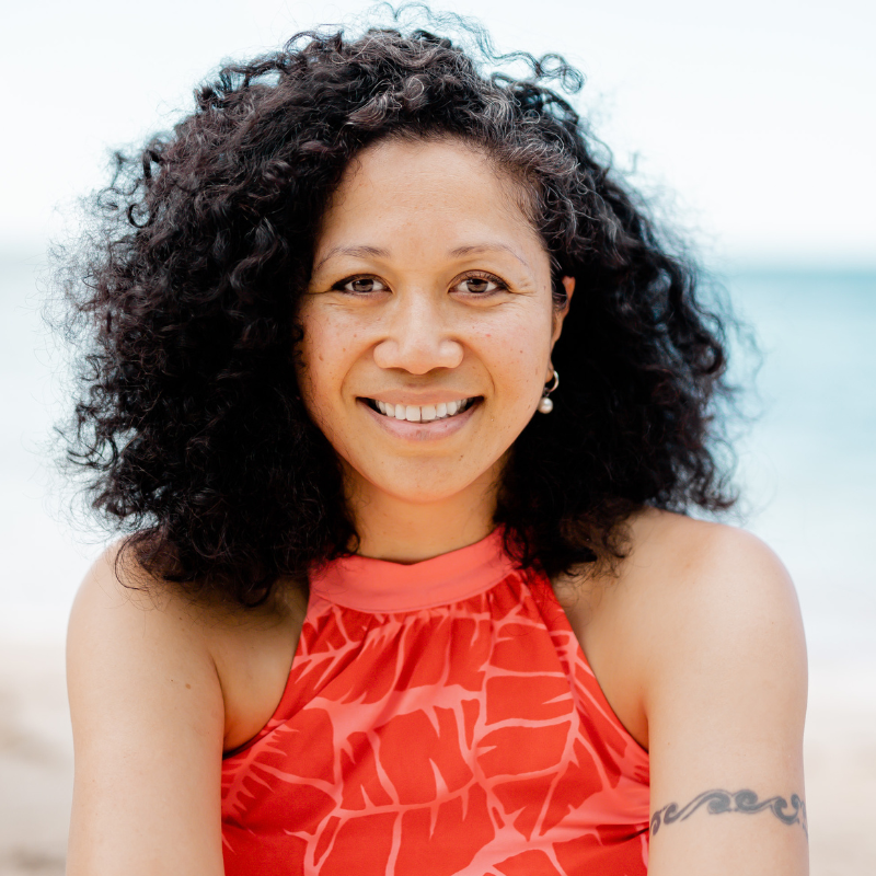 Samoan woman with dark curly hair, smiling and wearing a pink halter neck top. She is sitting on the beach.
