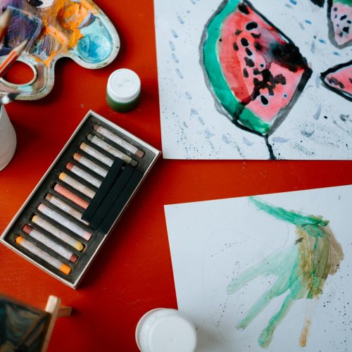 Creative art setup featuring watercolors and pastels on a red table, showcasing artistic expression.